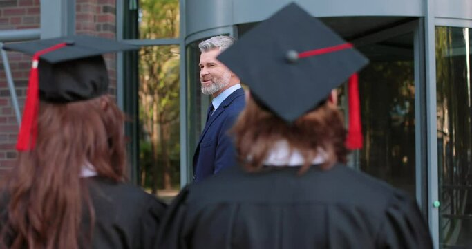 Full length view of the group of student people wearing graduation gowns and hats standing with their diplomas and celebrating graduation while listening speech of their teacher