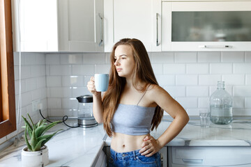Young happy woman drinking coffee or tea on the White scandinavian kitchen interior in the morning.wearing jeans and crop top.