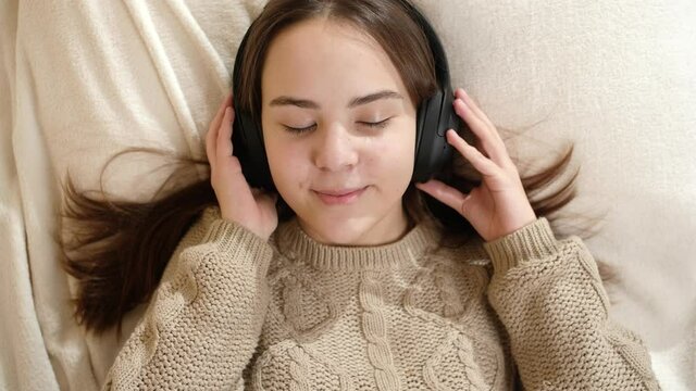 Top view of smiling teenage girl lying on bed ans listening music in headphones