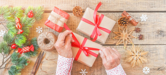 Senior woman packs gift for Christmas, top view. Wrinkled hands decorate surprise box with red ribbon, pine cones, holiday decoration, craft tools, vintage wooden table. Sharing family tradition.
