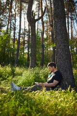 Handsome young man sitting in the forest on the grass reading a book