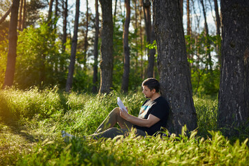 Naklejka na ściany i meble Handsome young man sitting in the forest on the grass reading a book
