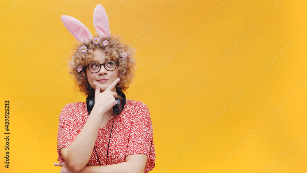 Wall mural Beautiful girl wearing cute bunny ears hairband and nerdy spectacles looking away from the camera. Thinking about something. Headphones around her neck. Isolated on a pink background studio.