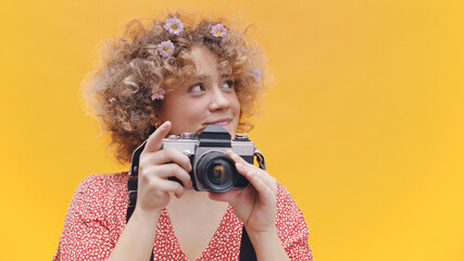 Attractive young girl holding camera in her hands. Dressed in casual pink top and wearing flowers in her hair. Isolated on yellow background studio. Concept of Photography. High quality photo. 