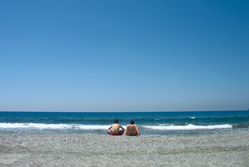 travel photography, two young guys are sitting on the seashore rear view on blue sky background closeup. Selective focus