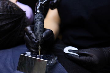hands of a tattoo artist wearing black gloves holding a tattoo machine in one hand and a cotton...