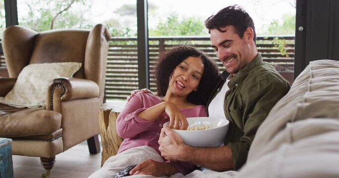 Smiling mixed race couple eating popcorn while watching a movie together at vacation home