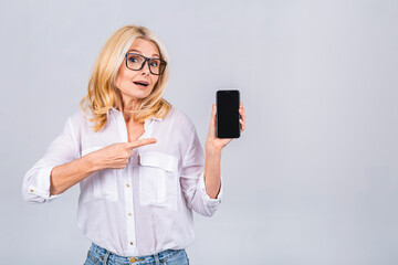 Surprised smiling happy senior mature woman in casual showing blank smartphone screen while looking at the camera isolated over white grey background. Using phone.