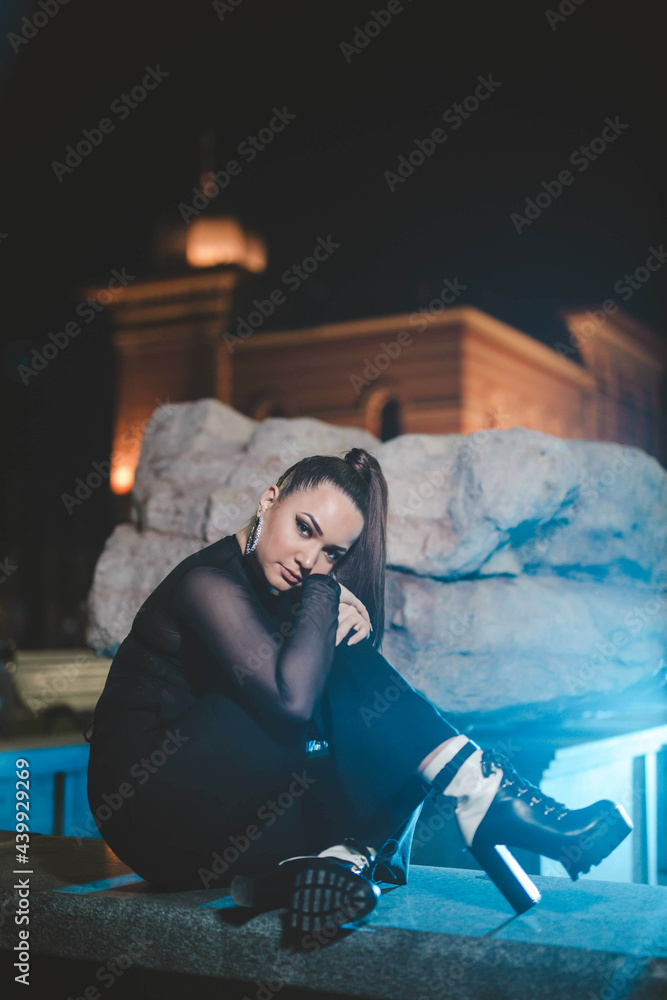 Poster Young attractive Caucasian girl sitting in front of a sculpture and posing outside in the evening