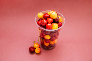 Ripe red and yellow cherries in a plastic cup on a pink background. Light snack.