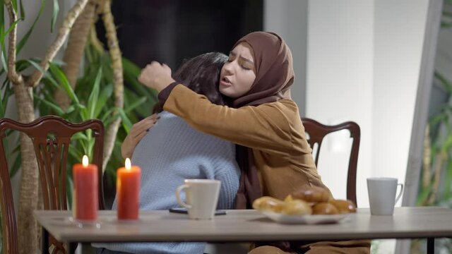 Middle Eastern young woman calming down stressed Caucasian friend complaining indoors in the evening. Supportive beautiful Muslim lady endorsing sad crying millennial indoors hugging