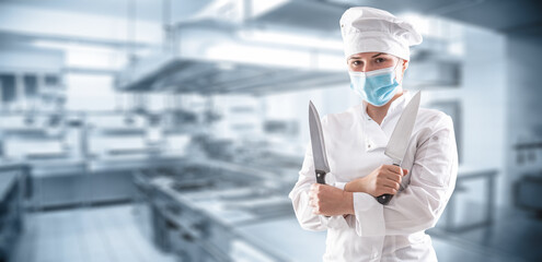 Panoramic banner of a chef with mask due to covid-19 pandemic  standing with crossed hands and holding two sharp knives in front of the restaurant kitchen