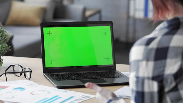 Over shoulder laptop screen view. Young woman has video call on laptop green screen. Female worker talking and listening during video conference with chroma key computer screen.