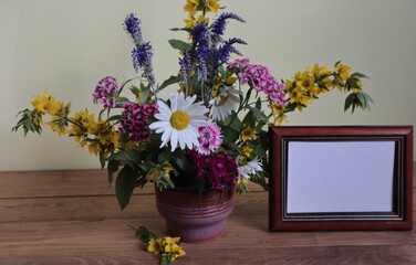 There is a bouquet of summer flowers in a vase and an empty photo frame on the table.
