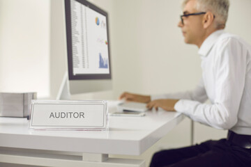 Closeup nameplate title sign plaque board that reads AUDITOR and senior man working on computer in background. Male worker sitting at office desk, looking at graphs and examining business data charts