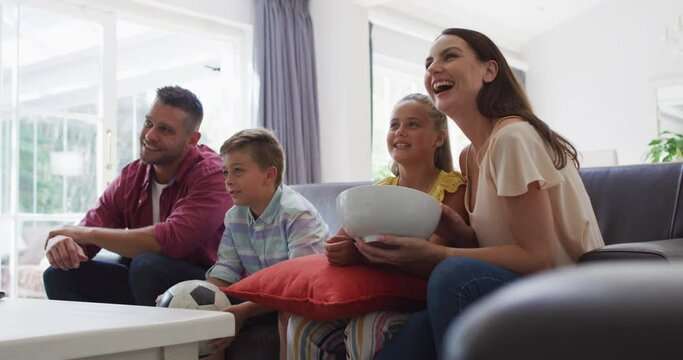 Excited caucasian parents, son and daughter on couch watching tv and cheering, son holding football