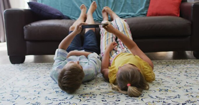 Happy caucasian brother and sister at home, lying on floor in living room using tablet together