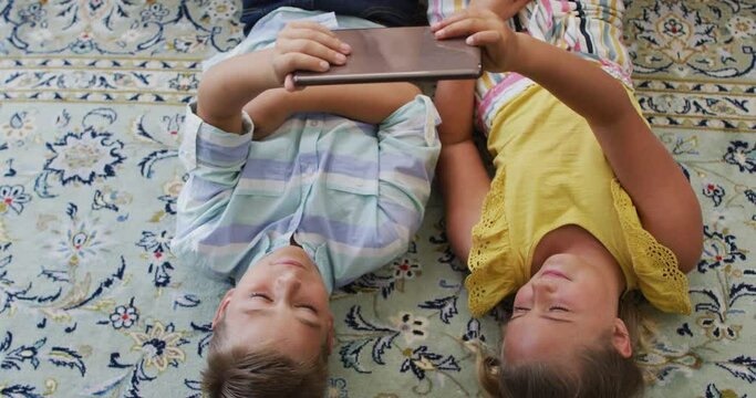 Happy caucasian brother and sister at home, lying on floor using tablet together and smiling