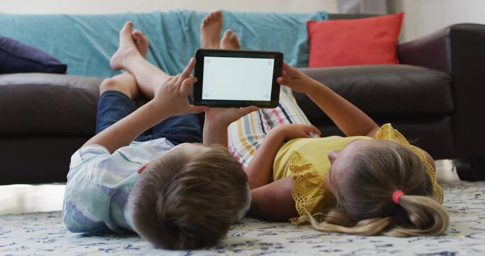 Happy caucasian brother and sister lying on floor in living room using tablet, copy space on screen