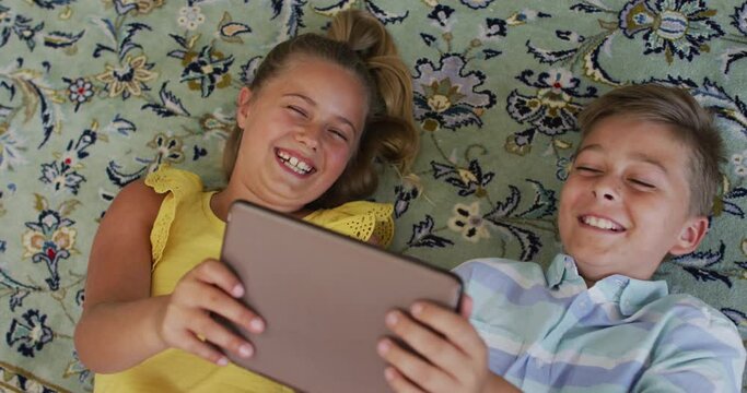 Happy caucasian brother and sister at home, lying on floor using tablet together and smiling