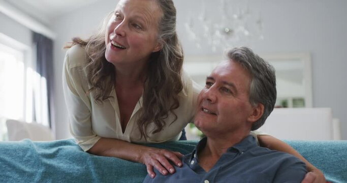 Happy senior caucasian couple watching tv in living room and talking, wife embracing sitting husband