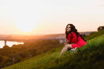 Beautiful young girl with long dark wavy hair sitting on a slope covered with green grass against the background of a summer sunset