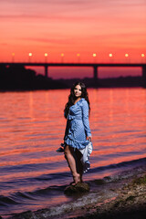 Beautiful young girl with long dark wavy hair standing at the bank of the river against the background of a bright sunset