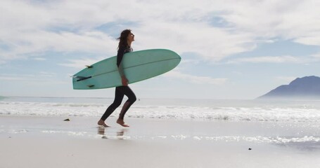 Happy mixed race woman running along beach by the sea carrying surfboard