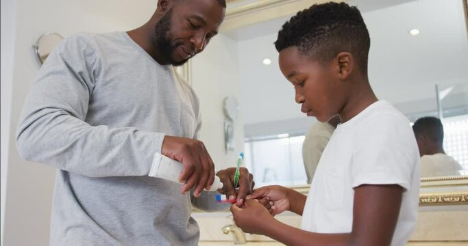 African american dad putting toothpaste on his son's brush in bathroom at home