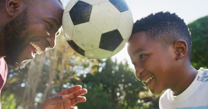 African american dad and son balancing the football together with their heads in the garden