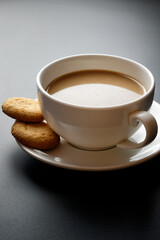 Latte in white coffee cup, with two cookies on the saucer. On black background in backlight.