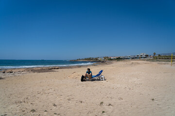 Fototapeta na wymiar Sad masked man sits on sun lounger in middle of empty beach on shores of Mediterranean Sea in Cyprus. Empty beach, no tourists. Lockdown and travel during coronavirus. Summer vacation at covid 19