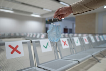 Travel essential concept, reopening flights during coronavirus quarantine. New normal subject. Social distancing chairs in airport and hand of traveler is holding medical mask. New rules in airport
