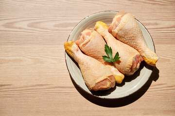 Top view of a raw chicken legs and parsley on a plate on a wooden table next to a window that lets...