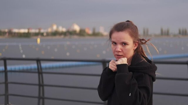 A young caucasian brunette woman delivers a series of boxing punches in a jump against the background of a river on the street in the afternoon. Sportswoman, boxing