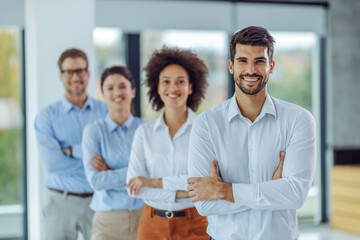Fototapeta premium Smiling multiracial group of business people standing with arms crossed and looking at camera while standing in office. Selective focus on man in foreground.