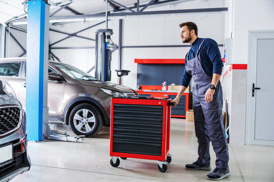 Mechanic pushing box with tools and preparing to fix a problem with car while walking in car salon.