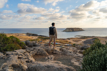Sea sunset view. Man with backpack on rocks with beautiful view of Yeronisos Island near coast of Agios Georgios Pegeias. Guy enjoying ocean horizon, panoramic sunset in cyprus on mediterranean sea