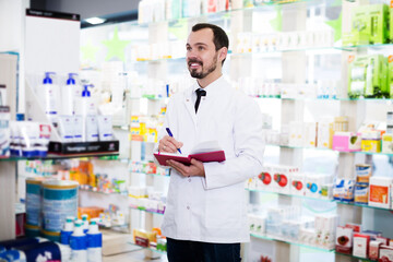 Young positive male pharmacist checking assortment of drugs in drugstore
