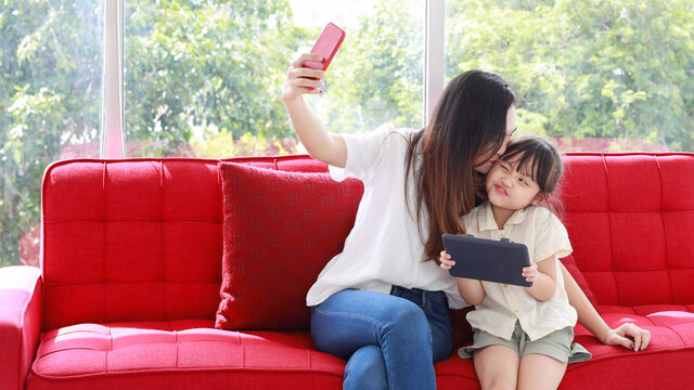 Asian long black hair mother wear white shirt and blue jeans sit smiling on big red sofa take selfie photo by red smartphone camera together with happy small daughter hold black tablet on her hands