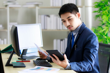 Successful young Asian businessman smiles happily while seeing good news on tablet in office. Business and success concept.