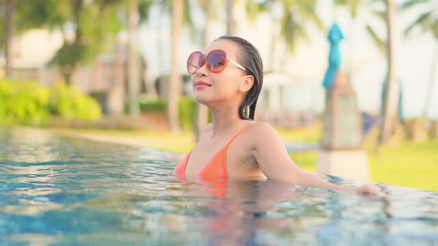 Woman wearing large sunglasses relaxing in infinity swimming pool of beautiful luxury tropical resort. Paradise vacation destination with young Asian woman in bright orange swimsuit tanning.