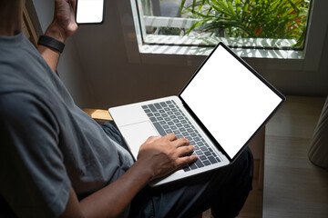 Casual man sitting in living room holding mobile phone and using laptop computer.