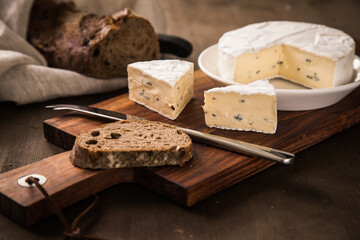 Loaf of soft blue cheese from cow milk on porcelain plate with walnut bread, knife, linen towel and dark brown wooden board as snack or dinner