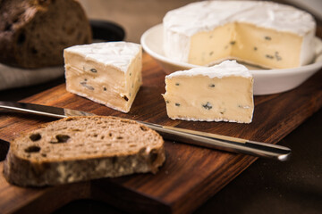 Loaf of soft blue cheese from cow milk on porcelain plate with walnut bread, knife, linen towel and dark brown wooden board as snack or dinner