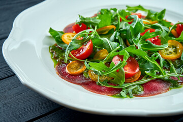 The traditional Italian appetizer salad is vitello tonnato. Thinly sliced beef with rucola, tomatoes and pesto, served in a white plate on a dark background