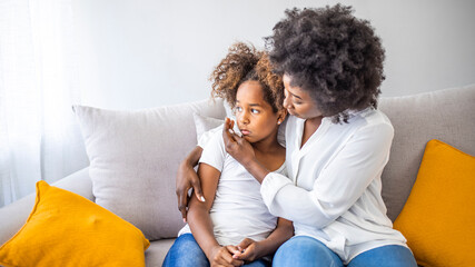 Black African mother embrace little preschool frustrated kid sitting on couch together at home. American loving mother supports disappointed daughter sympathizing, making peace after scolding concept