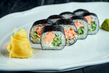 Appetizing Japanese futomak sushi roll with salmon, cucumber in a white plate on a black wooden background with ginger and wasabi. Japanese cuisine. Futomak roll
