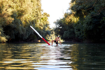 Fototapeta na wymiar Man making pivot or step back turn trick on stand-up paddle board (SUP) on the river near tress at suumer sunset. Extreme sport activity