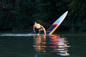 Back view on man making pivot or step back turn trick on stand-up paddle board (SUP) on the river near tress at suumer sunset. Extreme sport activity.